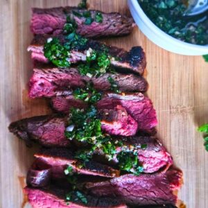 Marinated flank steak sliced and drizzled with chimichurri sauce on a cutting board with a side of chimichurri.