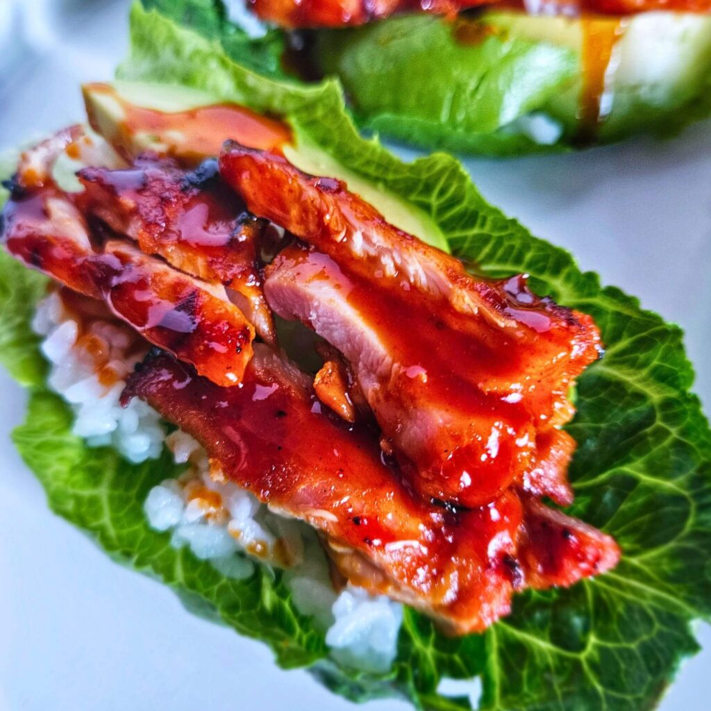 Close up image of grilled Korean BBQ chicken lettuce wraps layered with white rice, avocado, korean bbq chicken and drizzled with a spicy gochujang sauce.