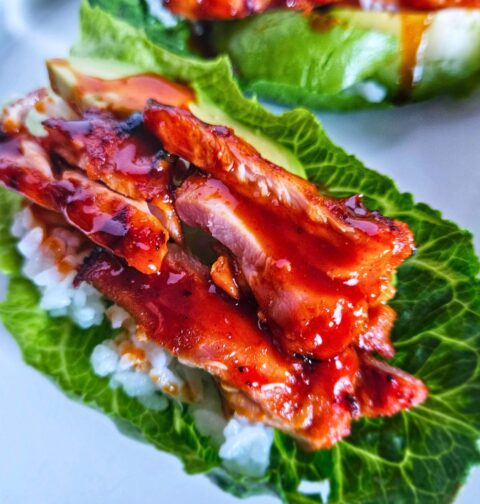 Close up image of grilled Korean BBQ chicken lettuce wraps layered with white rice, avocado, korean bbq chicken and drizzled with a spicy gochujang sauce.