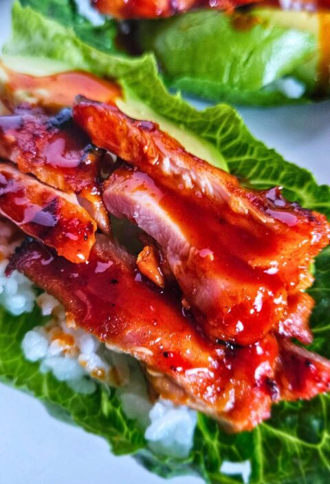 Close up image of grilled Korean BBQ chicken lettuce wraps layered with white rice, avocado, korean bbq chicken and drizzled with a spicy gochujang sauce.