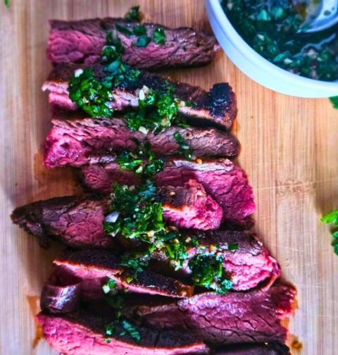 Close up image of marinated flank steak sliced and drizzled with chimichurri sauce on a cutting board with a side of chimichurri.