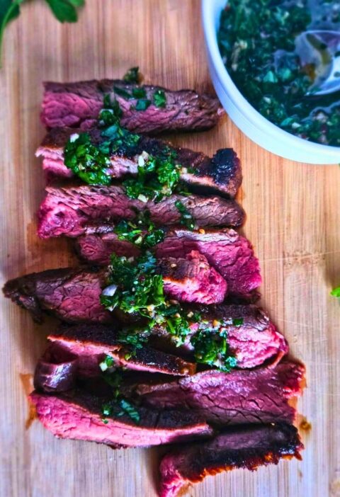 Close up image of marinated flank steak sliced and drizzled with chimichurri sauce on a cutting board with a side of chimichurri.
