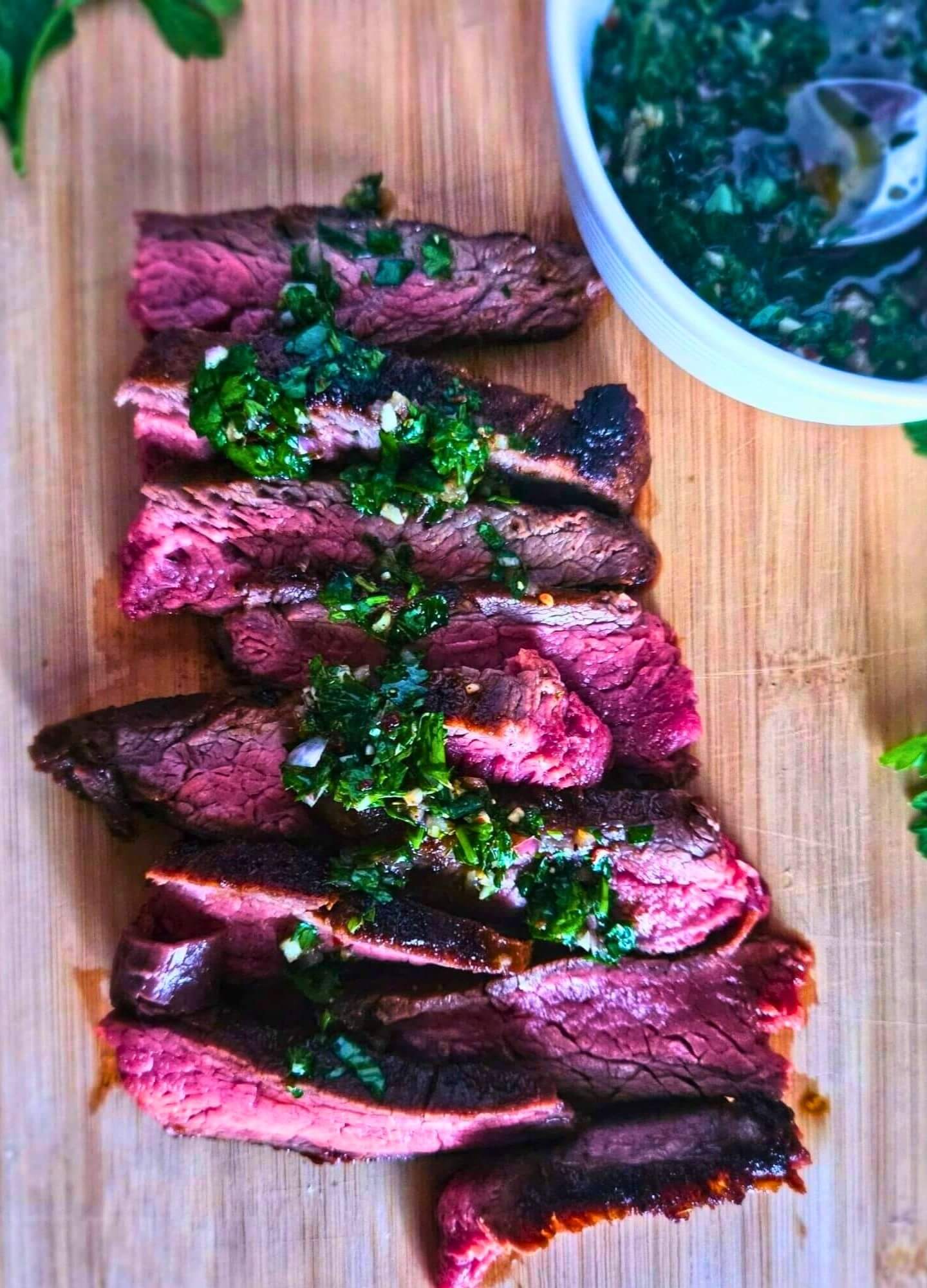 Photo of marinated flank steak sliced on a cutting board with chimichurri sauce drizzled on top. 
