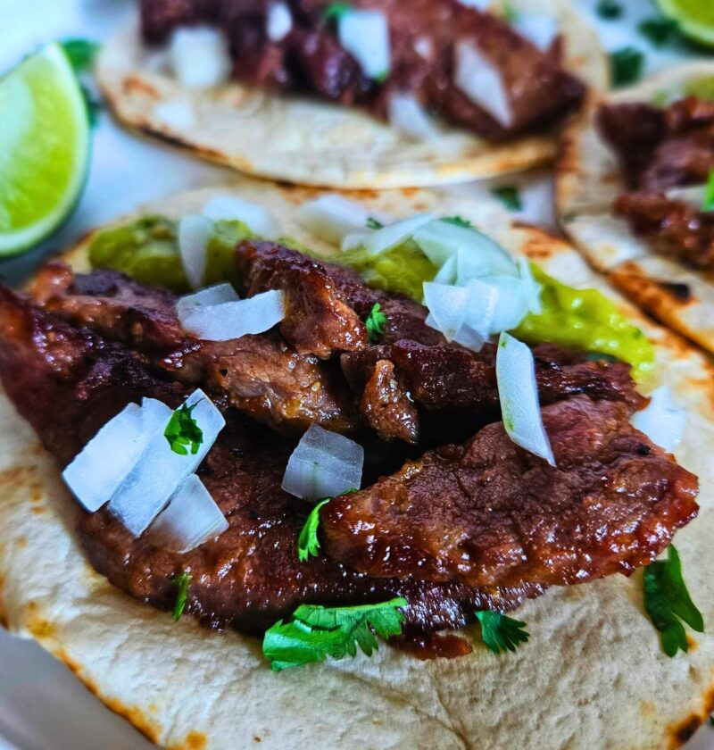 Close up image of steak tacos with marinated flank steak, chopped white onions and a creamy avocado tomatillo salsa.