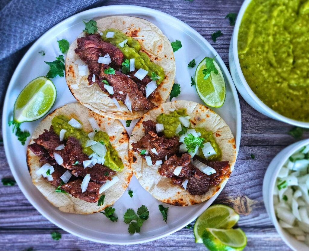 Steak tacos 8 Steak tacos with chopped white onions and avocado tomatillo salsa on a plate served with limes and cilantrol.