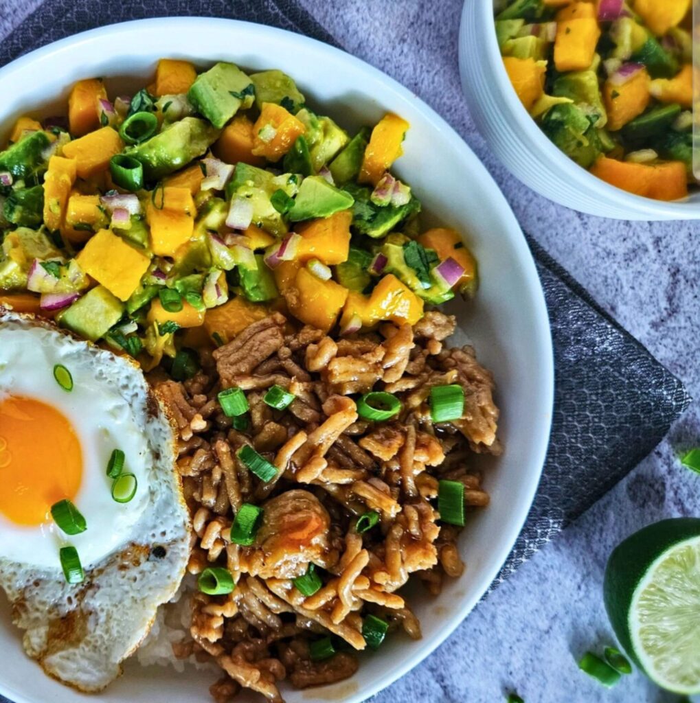 Image of teriyaki chicken rice bowl with avocado mango salsa over white rice with a fried egg.