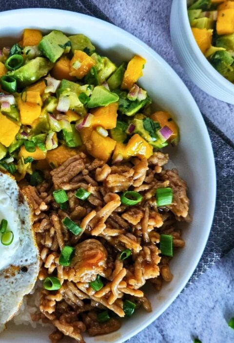 Image of teriyaki chicken rice bowl with avocado mango salsa over white rice with a fried egg.