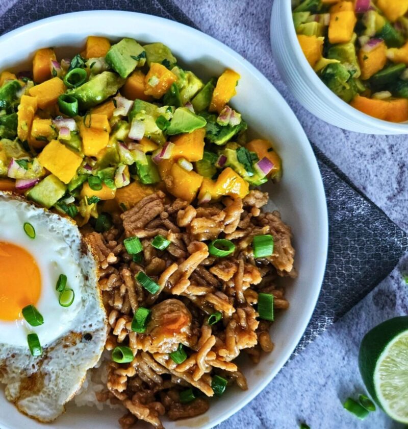Image of teriyaki chicken rice bowl with avocado mango salsa over white rice with a fried egg.