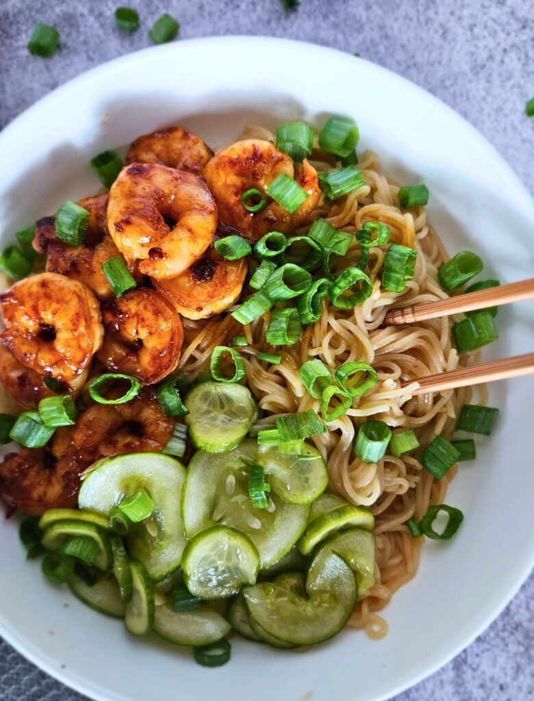 Image of peanut noodles with chili shrimp and pickled cucumbers in a bowl with chopsticks.