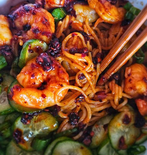 Image of peanut noodles with chili shrimp and pickled cucumbers in a bowl with chopstick being eaten.