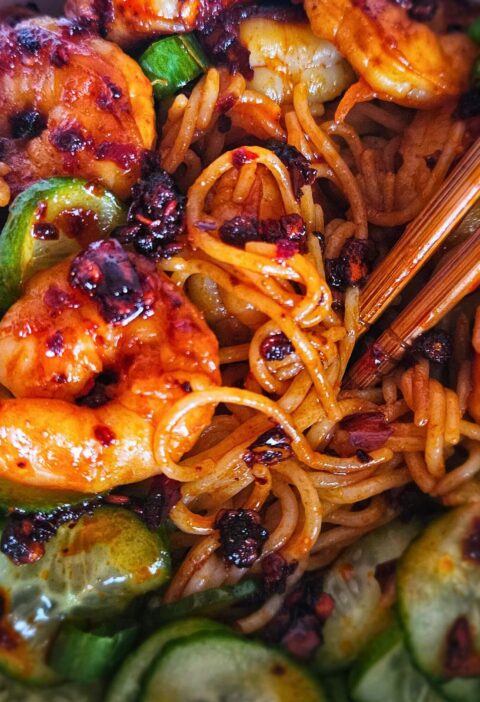 Image of peanut noodles with chili shrimp and pickled cucumbers in a bowl with chopstick being eaten.