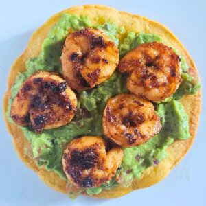 Shrimp tostada being assemble with homemade tostada, guacamole, and blackened shrimp.