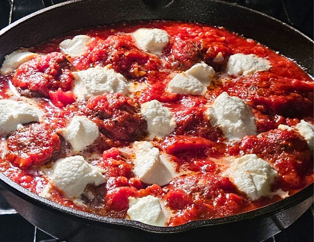 Ricotta meatballs in homemade marinara sauce in a cast iron skillet baking in the oven with dollops of ricotta cheese on top.
