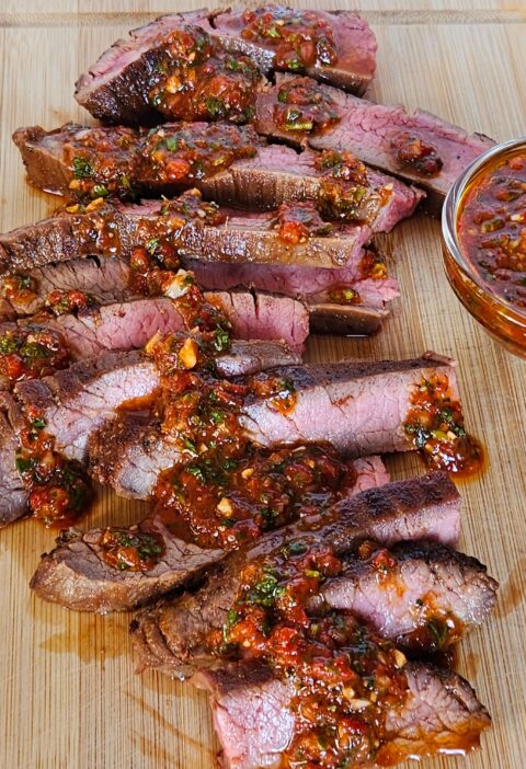 Image of cast iron flank steak sliced on cutting board drizzle with red chimichurri.