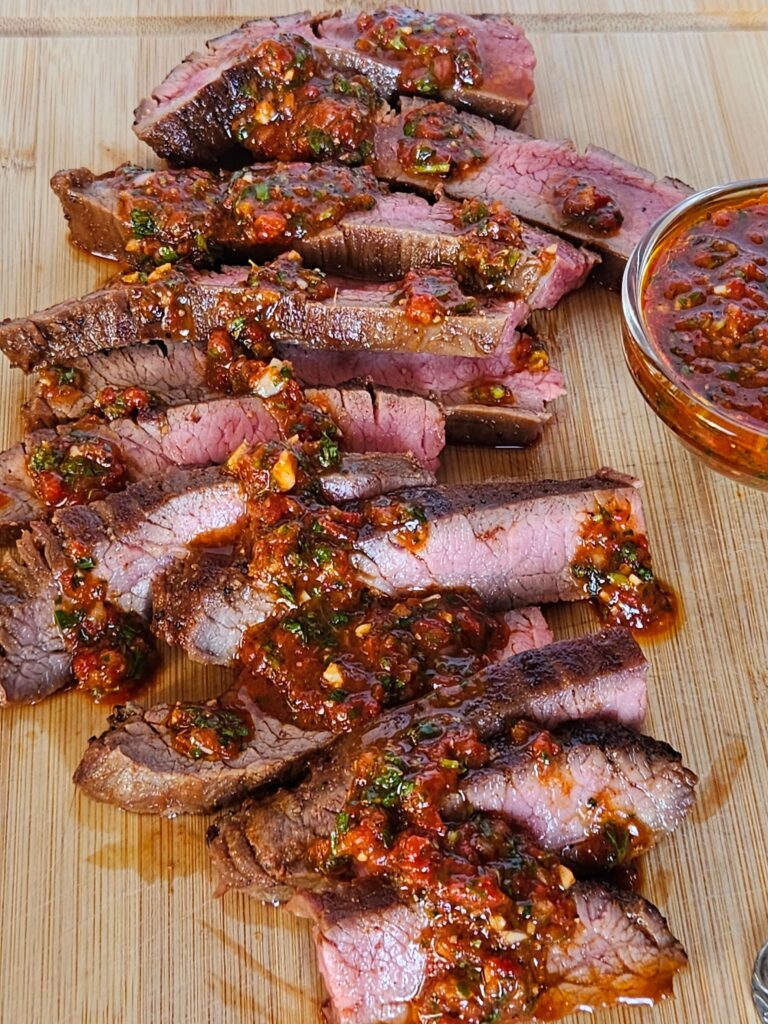 Image of cast iron flank steak sliced on cutting board drizzle with red chimichurri.