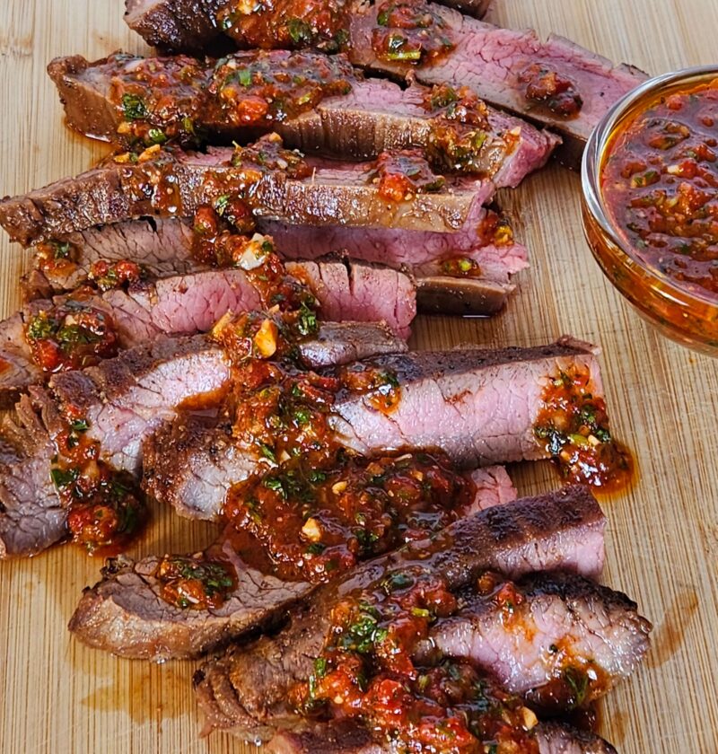 Image of cast iron flank steak sliced on cutting board drizzle with red chimichurri.