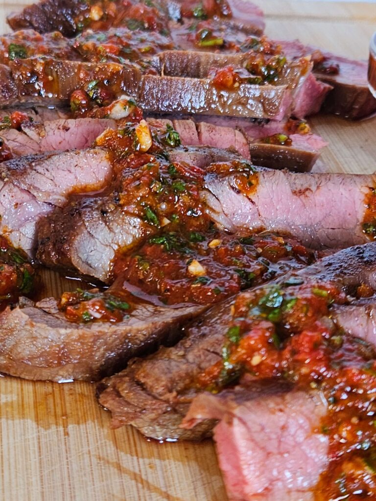 Close up image of cast iron flank steak sliced on a cutting board with red chimichurri drizzled on top.