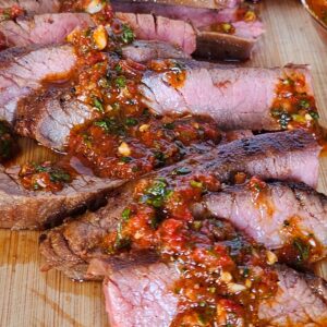 Close up image of cast iron flank steak sliced on a cutting board with red chimichurri.