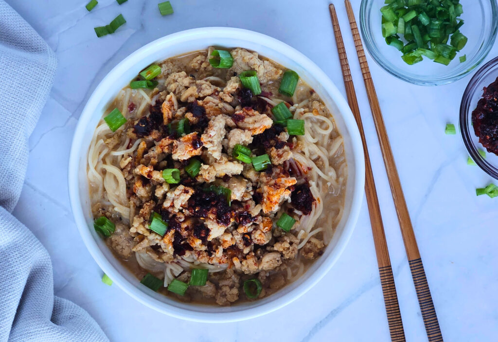 Ramen 2 Image of ramen being served in red chili coconut broth and topped with ground chicken, chili crispy and green onions.