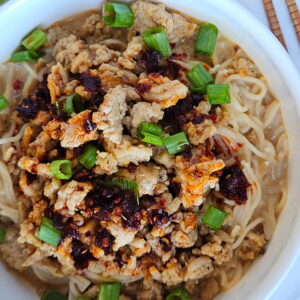 Image of ramen in a red chili coconut broth and topped with ground chicken, chili crisp and green onions.
