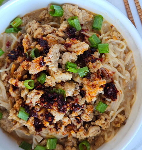 Image of ramen in a red chili coconut broth and topped with ground chicken, chili crisp and green onions.
