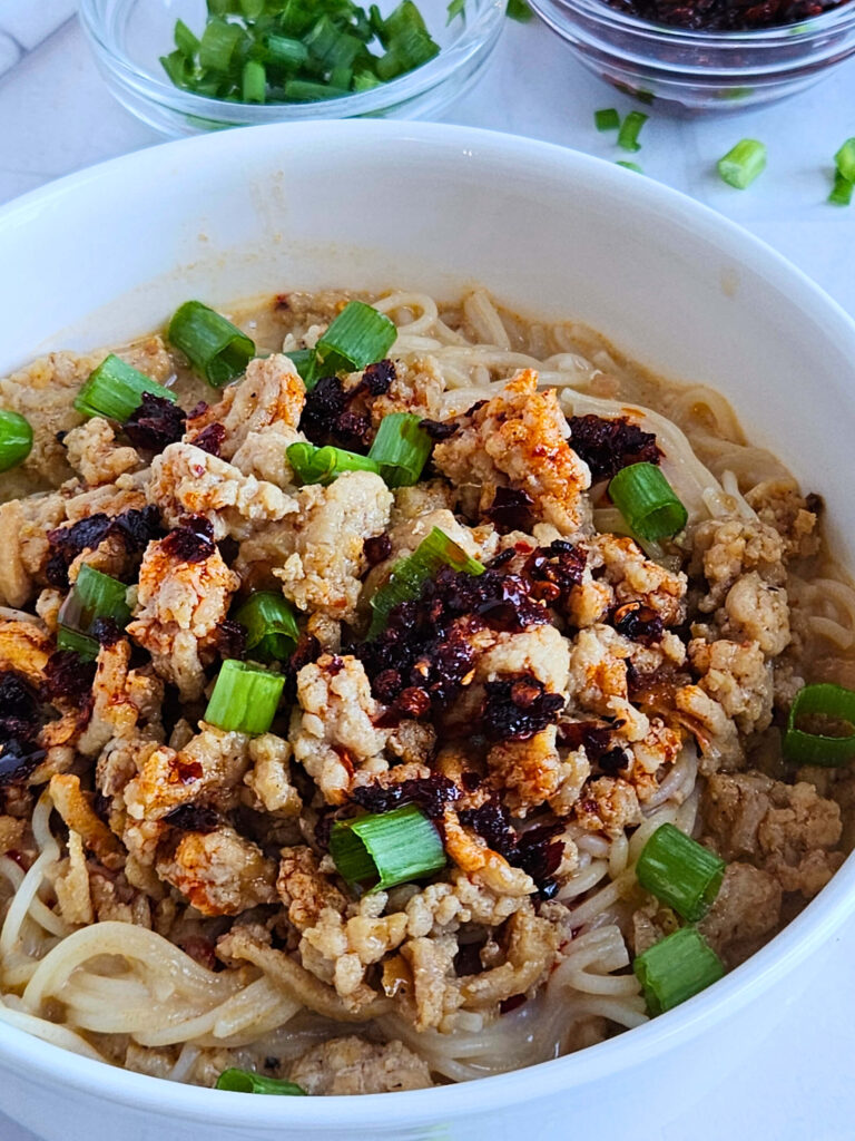 Ramen 8 Close up image of ramen in a red chili coconut broth topped with ground chicken, chili crisp and green onions.